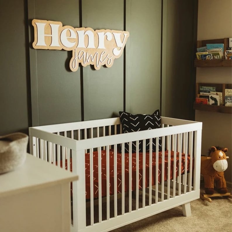 May include: A white crib with a red and white patterned sheet and a black and white pillow. The crib is in a room with a green wall and a wooden shelf with books. A wooden name sign with the names "Henry James" is on the wall above the crib.