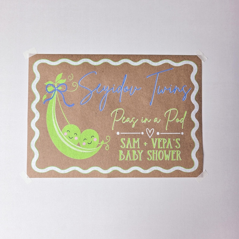 May include: A rectangular sign with a brown background and a white wavy border. The sign features a green pea pod with two smiling peas and the text "Seyidov Twins Peas in a Pod SAM + VEPA'S BABY SHOWER" in blue and green.
