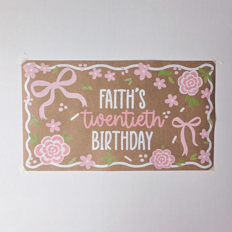 May include: A rectangular brown paper sign featuring the white and pink text "FAITH'S twentieth BIRTHDAY". The design includes a white scalloped border, light pink bows, and pink floral illustrations with small green leaves scattered throughout.