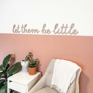 May include: A wall-mounted wooden sign featuring the phrase let them be little in a cursive script font. The letters are cut from a light-colored wood and are mounted horizontally on a wall painted with a white upper section and a dusty rose lower section.