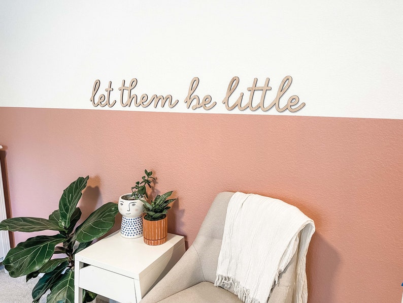 May include: A wall-mounted wooden sign featuring the phrase let them be little in a cursive script font. The letters are cut from a light-colored wood and are mounted horizontally on a wall painted with a white upper section and a dusty rose lower section.