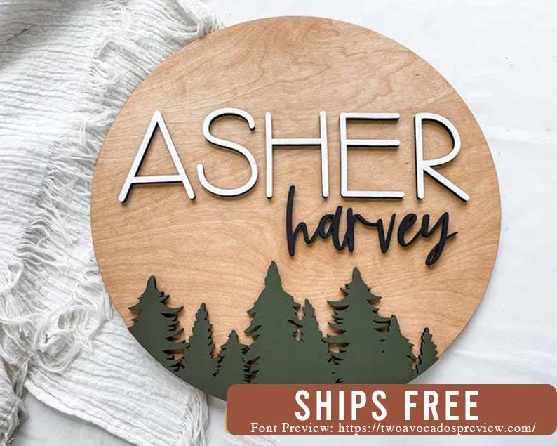 Op de afbeelding: Een rond houten bord met de namen "Asher Harvey" in witte en zwarte letters. Het bord heeft een bos sc&egrave;ne met groene bomen aan de onderkant.