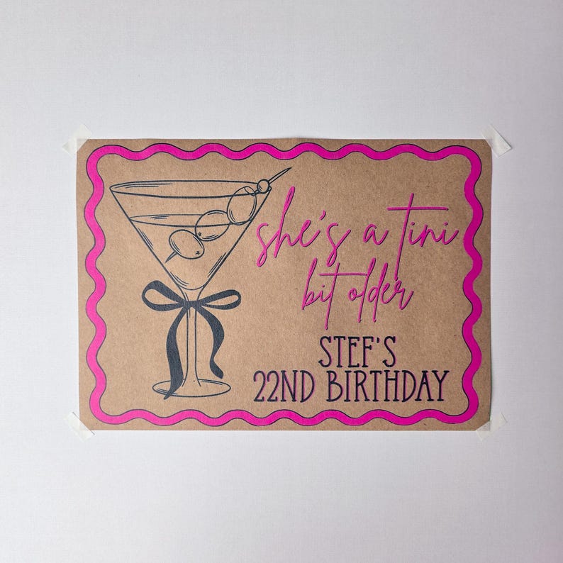 May include: A rectangular sign on brown paper with a bright pink scalloped border. It features a black line drawing of a martini glass with olives and a bow, with the pink text "she's a tini bit older" and black text "STEF'S 22ND BIRTHDAY".
