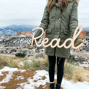 May include: A light-colored wooden cutout sign featuring the word Read in a continuous cursive script font. The letters are connected in a single piece, showcasing a smooth, natural wood grain finish with clean, laser-cut edges throughout the design.