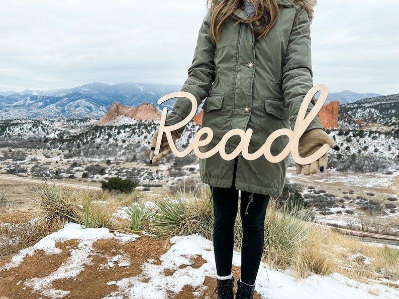 May include: A light-colored wooden cutout sign featuring the word Read in a continuous cursive script font. The letters are connected in a single piece, showcasing a smooth, natural wood grain finish with clean, laser-cut edges throughout the design.