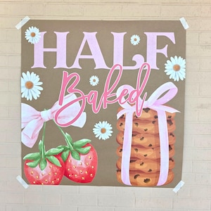 May include: A brown poster with the words "HALF BAKED" in pink, featuring illustrations of strawberries, daisies, and a stack of chocolate chip cookies tied with a pink ribbon. The poster is taped to a brick wall.