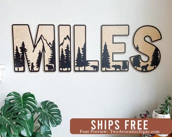 May include: A set of five large wooden letters spelling the name MILES, featuring a natural light wood finish with black cut-out silhouettes of mountain ranges, pine trees, and bears integrated into the design of each individual letter.