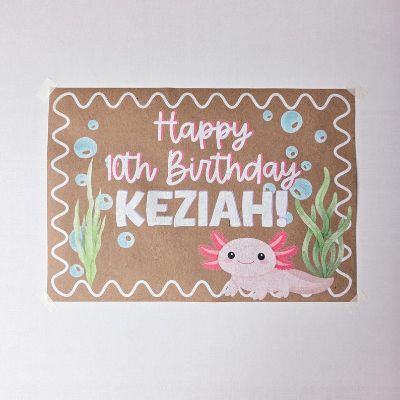 May include: A brown paper birthday card with the text "Happy 10th Birthday KEZIAH!" in pink and white. The card features a cartoon axolotl, bubbles, and seaweed illustrations. The card is decorated with a white wavy border.