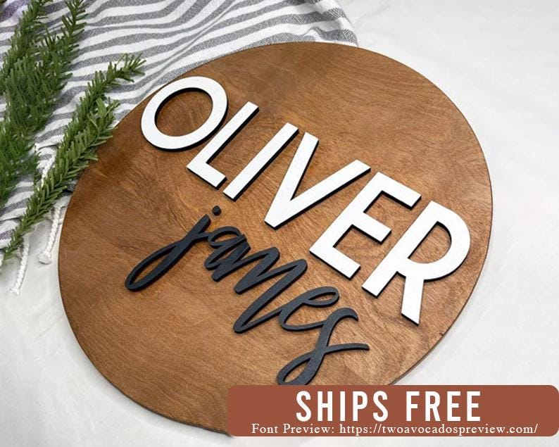 May include: A circular wooden sign with a smooth, medium-brown stained finish. The name OLIVER is displayed in large, white, uppercase sans-serif letters, while the name james is written below in smaller, black, lowercase script lettering.