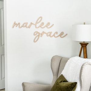 Pode incluir: Uma parede branca com uma placa de madeira que diz "marlee grace" em escrita cursiva. Um abajur branco com uma base de trip&eacute; de madeira est&aacute; em uma poltrona branca com um cobertor branco e uma almofada verde.