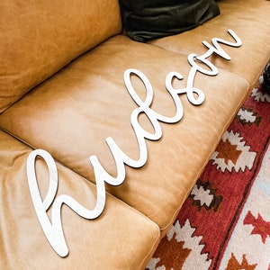 May include: A large, white, cursive-style wooden cutout sign displaying the name Hudson in a continuous, flowing script font. The letters are connected and feature a smooth, matte finish, designed as a decorative wall or room accent piece.