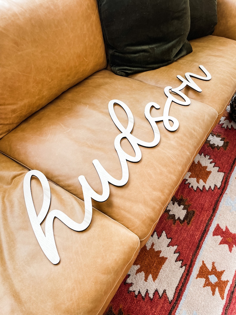 May include: A large, white, cursive-style wooden cutout sign displaying the name Hudson in a continuous, flowing script font. The letters are connected and feature a smooth, matte finish, designed as a decorative wall or room accent piece.