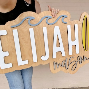 Puede incluir: Un letrero de madera con el nombre "Elijah" y la palabra "mason" escrita en cursiva. El letrero est&aacute; decorado con olas azules y una tabla de surf amarilla.