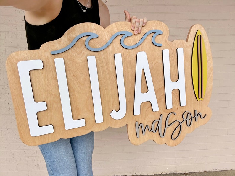 Puede incluir: Un letrero de madera con el nombre "Elijah" y la palabra "mason" escrita en cursiva. El letrero est&aacute; decorado con olas azules y una tabla de surf amarilla.