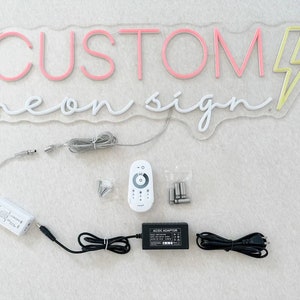 K&ouml;nnte beinhalten: Ein pinkfarbenes und gelbes Neon-Schild mit der Aufschrift "CUSTOM neon sign" und einem Blitz. Das Schild befindet sich auf einer wei&szlig;en Oberfl&auml;che mit einer wei&szlig;en Fernbedienung, einem Netzteil und einem Kabel.