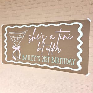 May include: A brown banner with a white wavy border and the text "she's a tini bit older" in pink and white script. A martini glass illustration is on the left, and "BAILEY'S 21ST BIRTHDAY" is in green.