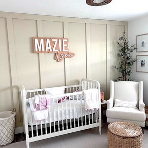 K&ouml;nnte beinhalten: Ein wei&szlig;es Babybett mit einer wei&szlig;en Matratze und Bettw&auml;sche, ein wei&szlig;er Sessel mit einem rosa Kissen, ein Weidenkorb und ein Holzschild mit dem Namen "Mazie Lynn" an einer beigefarbenen Wand.