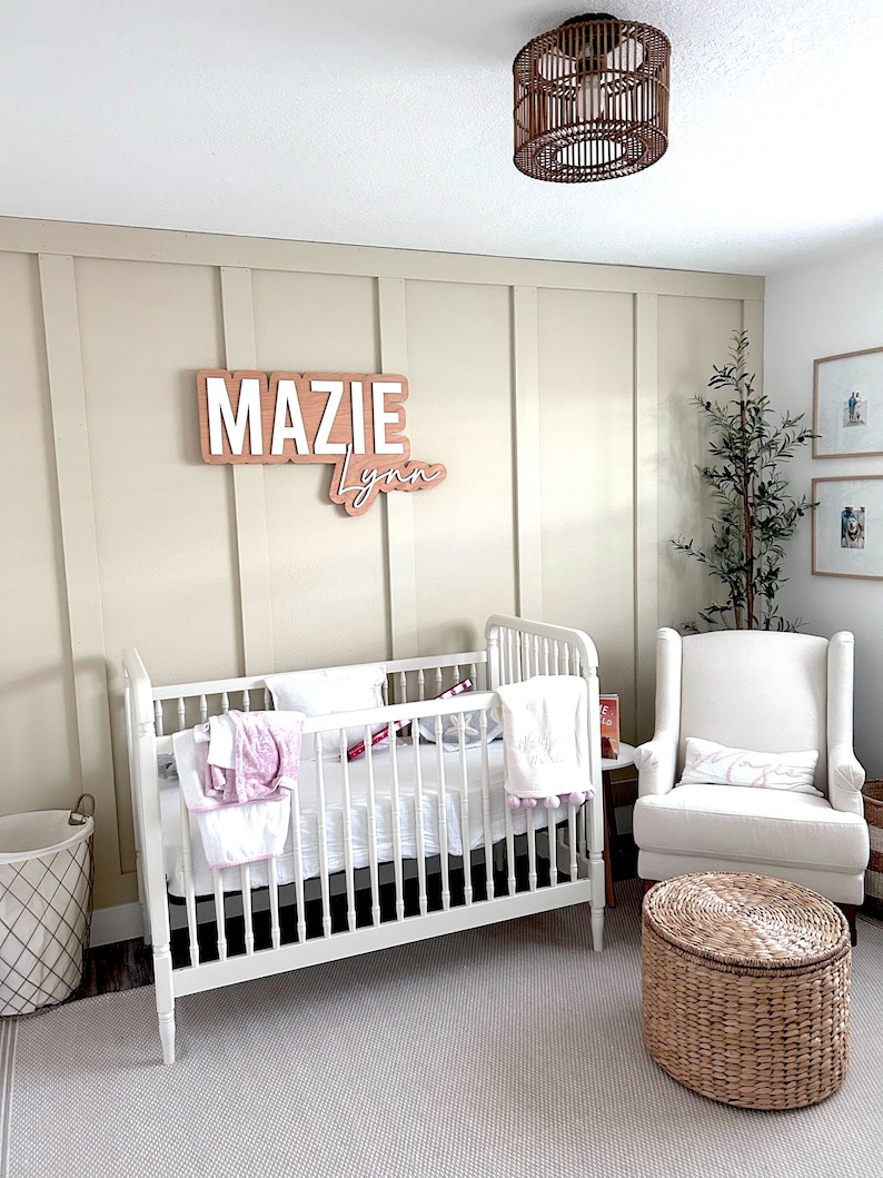 K&ouml;nnte beinhalten: Ein wei&szlig;es Babybett mit einer wei&szlig;en Matratze und Bettw&auml;sche, ein wei&szlig;er Sessel mit einem rosa Kissen, ein Weidenkorb und ein Holzschild mit dem Namen "Mazie Lynn" an einer beigefarbenen Wand.