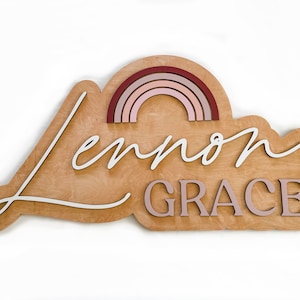 May include: Wooden sign with a cloud shape, featuring the names "Lennon Grace" in white lettering and a rainbow made of layered wood in shades of pink and red.