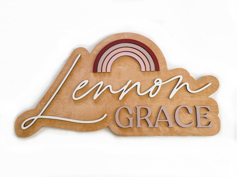 May include: Wooden sign with a cloud shape, featuring the names "Lennon Grace" in white lettering and a rainbow made of layered wood in shades of pink and red.