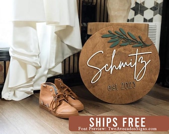 May include: A round wooden sign featuring the name Schmitz in white script lettering, the text est 2023 in dark brown, and a dark green leafy branch cutout at the top. The sign is displayed next to a pair of tan high-top sneakers and white fabric.