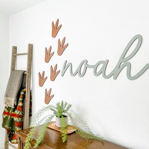 Pu&ograve; includere: Un cartello in legno grigio chiaro con il nome "noah" scritto in corsivo. Il cartello &egrave; montato su una parete bianca con impronte di dinosauro marroni.