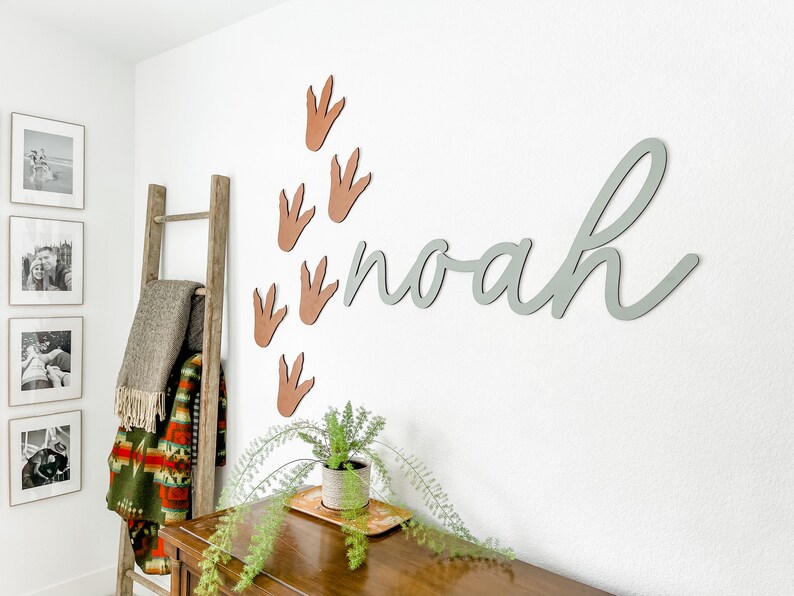 Pu&ograve; includere: Un cartello in legno grigio chiaro con il nome "noah" scritto in corsivo. Il cartello &egrave; montato su una parete bianca con impronte di dinosauro marroni.