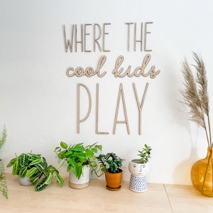 May include: A wall-mounted wooden sign featuring the text WHERE THE cool kids PLAY in a combination of block and script lettering. The sign is made of light-colored wood and is displayed on a plain wall above a collection of various potted plants.