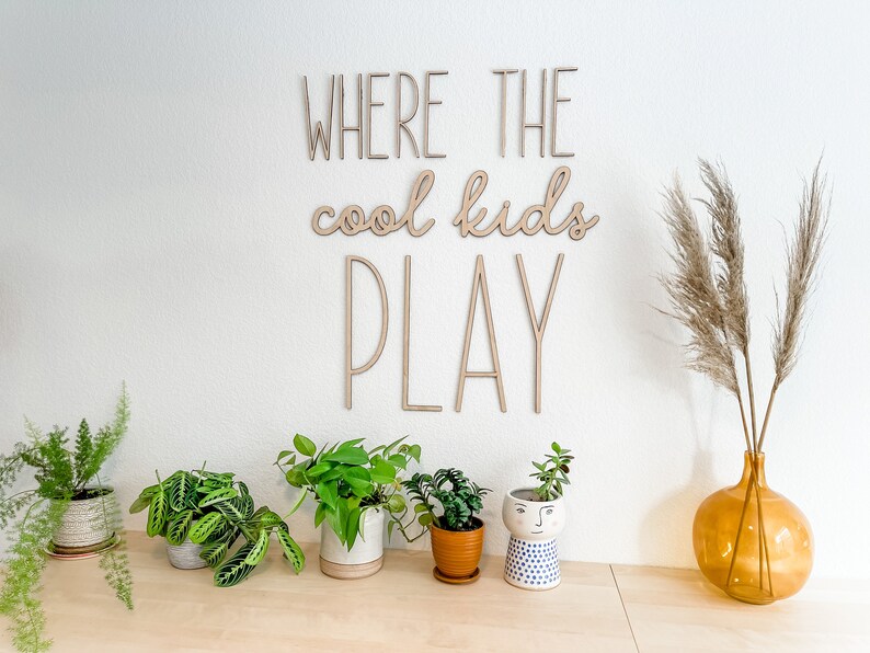 May include: A wall-mounted wooden sign featuring the text WHERE THE cool kids PLAY in a combination of block and script lettering. The sign is made of light-colored wood and is displayed on a plain wall above a collection of various potted plants.