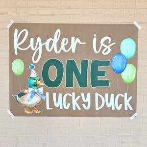 May include: A brown paper sign with the text "Ryder is ONE LUCKY DUCK" in white and green. A cartoon duck wearing a party hat and bow tie is on the left. Balloons in green and blue are on the right.
