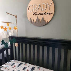Op de afbeelding: Een houten bord voor een kinderkamer met de namen "Grady Matthew" in witte en zwarte letters, met een bos-silhouetontwerp in bruin. Het bord hangt aan een groene muur boven een zwart ledikant met een wit en groen bos-thema hoeslaken.