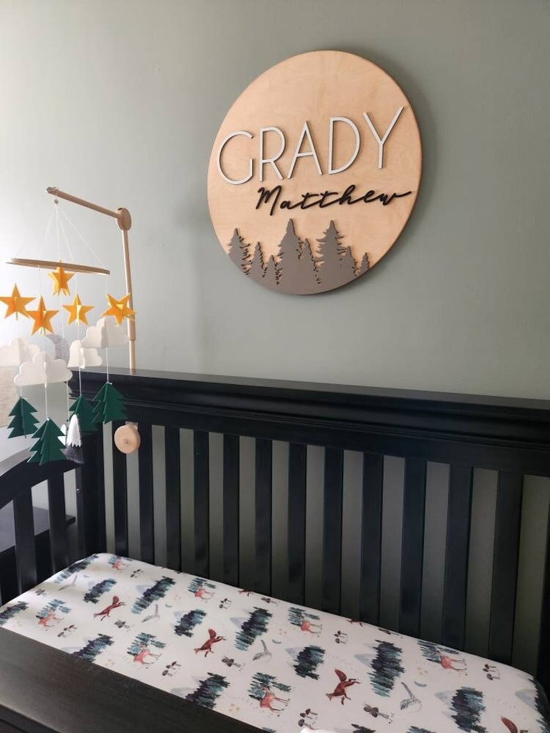 Op de afbeelding: Een houten bord voor een kinderkamer met de namen "Grady Matthew" in witte en zwarte letters, met een bos-silhouetontwerp in bruin. Het bord hangt aan een groene muur boven een zwart ledikant met een wit en groen bos-thema hoeslaken.