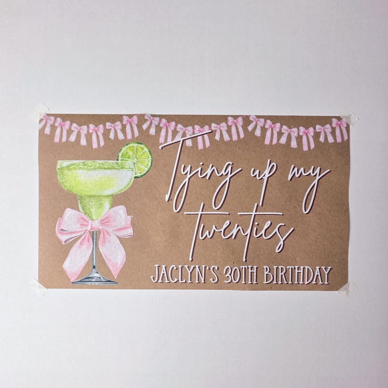 May include: A rectangular sign on brown paper featuring a green margarita glass with a lime slice and a pink bow. Above is a pink bow garland. The text reads: Tying up my Twenties, JACLYN'S 30TH BIRTHDAY in white script and block lettering.
