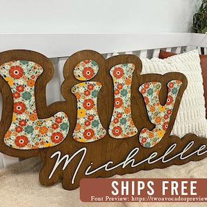May include: A wooden wall sign featuring the name Lily in large, block-style letters with a floral pattern of red, orange, and teal flowers on a cream background. Below, the name Michelle is written in white cursive script on the wood base.
