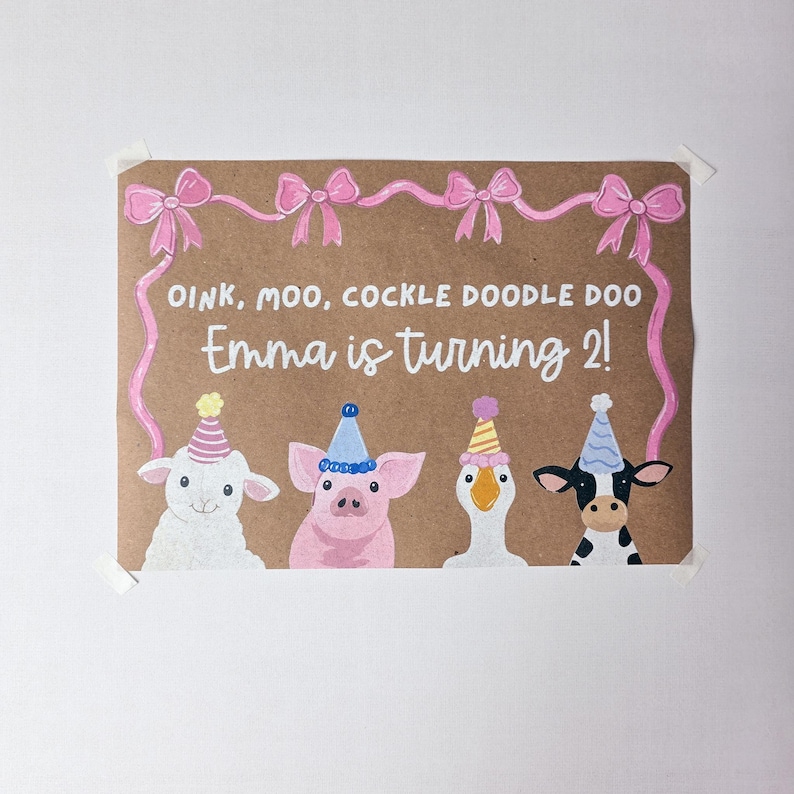 May include: A brown paper sign featuring a pink ribbon border and the text OINK, MOO, COCKLE DOODLE DOO Emma is turning 2! Below the text are illustrations of a white lamb, a pink pig, a white goose, and a black and white cow, all wearing party hats.