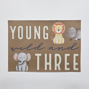 May include: A rectangular brown card with the words "YOUNG wild and THREE" in white and blue lettering. The card features illustrations of a gray elephant, a yellow lion, and a gray hippo.