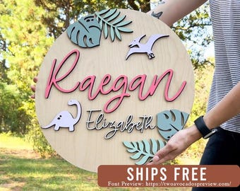 May include: A circular wooden sign featuring the name Raegan in large pink script and Elizabeth in black script. The design includes light green monstera and palm leaves, a light purple dinosaur, and a light purple pterodactyl cutout.