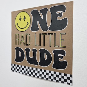 May include: A brown card with the text "ONE RAD LITTLE DUDE" in black and yellow lettering. A yellow smiley face with lightning bolt eyes is on the left. A black and white checkered pattern is at the bottom.