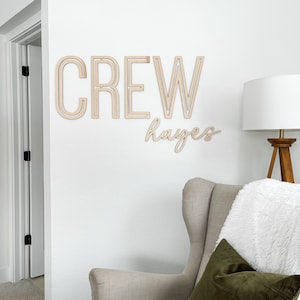Pode incluir: Decora&ccedil;&atilde;o de parede de madeira com a palavra "CREW" em letras grandes e o nome "Hayes" escrito em cursiva abaixo. A decora&ccedil;&atilde;o &eacute; exibida em uma parede branca acima de uma poltrona bege com uma manta branca e uma almofada verde.