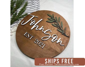 May include: A circular wooden sign with a dark brown stained finish. The sign features the white 3D lettering "Johnson" in a cursive script and "EST 2021" in a block font. An olive green 3D leaf sprig is attached to the upper right side.