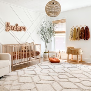 K&ouml;nnte beinhalten: Ein Kinderzimmer mit einem wei&szlig;en Babybett, einem wei&szlig;en Teppich mit geometrischem Muster, einer H&auml;ngeleuchte aus Weidengeflecht, einem Topfbaum mit Olivenbaum und einem Holzschild mit der Aufschrift "Beckham".