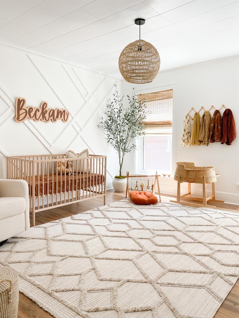 K&ouml;nnte beinhalten: Ein Kinderzimmer mit einem wei&szlig;en Babybett, einem wei&szlig;en Teppich mit geometrischem Muster, einer H&auml;ngeleuchte aus Weidengeflecht, einem Topfbaum mit Olivenbaum und einem Holzschild mit der Aufschrift "Beckham".