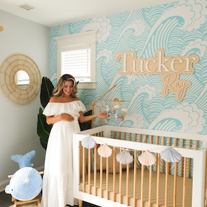 K&ouml;nnte beinhalten: Ein wei&szlig;es Babybett mit einem Holzrahmen und einer wei&szlig;en Matratze. Ein blaues und wei&szlig;es Mobile h&auml;ngt &uuml;ber dem Bett. Ein wei&szlig;er Teppich mit geometrischem Muster liegt auf dem Boden. Ein blaues und wei&szlig;es Schaukelpferd in Form eines Wals steht im Vordergrund. Die Wand hinter dem Bett ist hellblau gestrichen und zeigt ein wei&szlig;es Wellenmuster. Ein Holzschild mit dem Namen "Tucker Ray" befindet sich an der Wand.