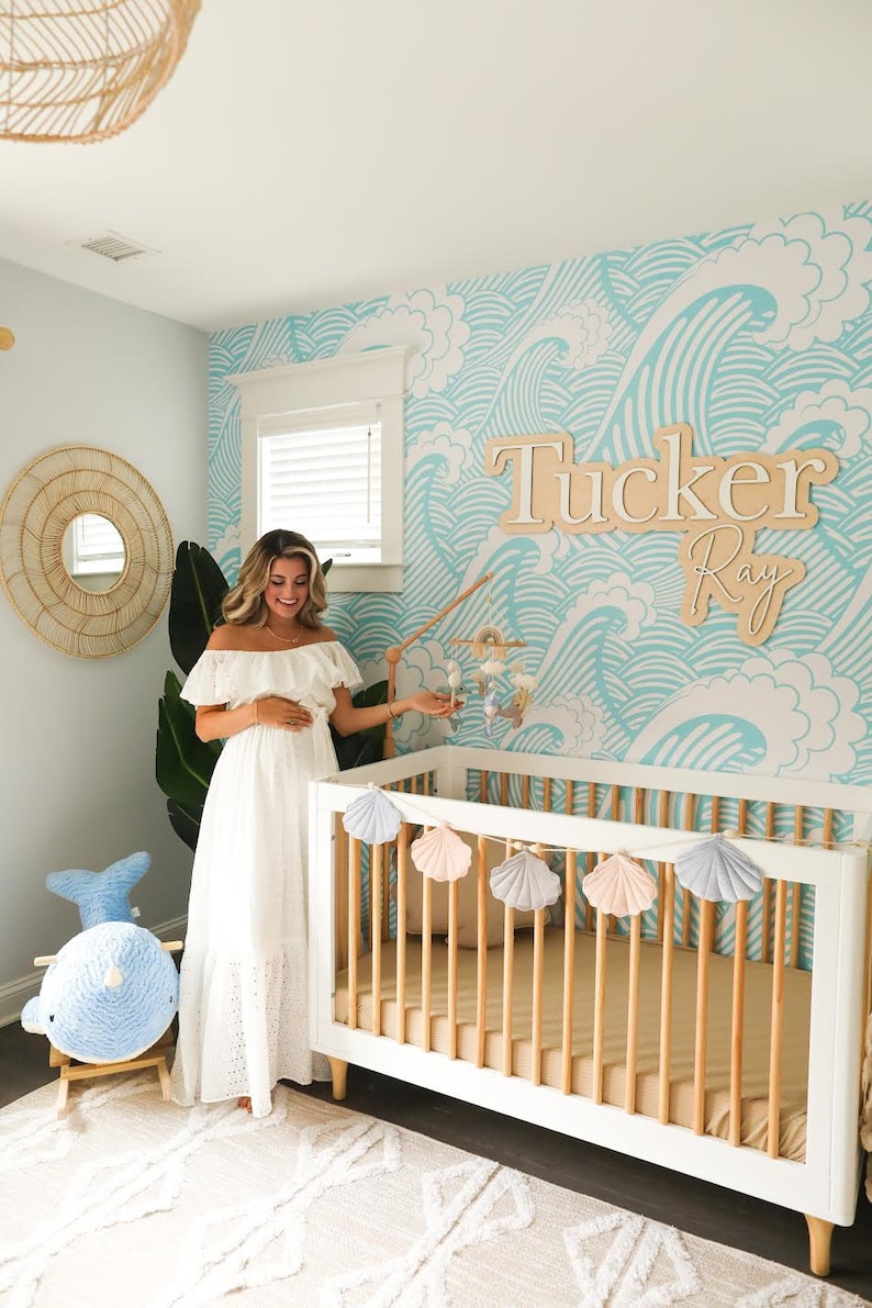 K&ouml;nnte beinhalten: Ein wei&szlig;es Babybett mit einem Holzrahmen und einer wei&szlig;en Matratze. Ein blaues und wei&szlig;es Mobile h&auml;ngt &uuml;ber dem Bett. Ein wei&szlig;er Teppich mit geometrischem Muster liegt auf dem Boden. Ein blaues und wei&szlig;es Schaukelpferd in Form eines Wals steht im Vordergrund. Die Wand hinter dem Bett ist hellblau gestrichen und zeigt ein wei&szlig;es Wellenmuster. Ein Holzschild mit dem Namen "Tucker Ray" befindet sich an der Wand.
