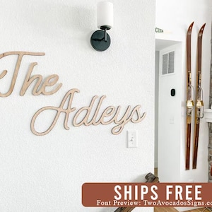 May include: Wooden wall decor with the text "The Adleys" in a cursive font. The sign is mounted on a white wall with a light fixture above it.