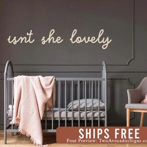 May include: A wall-mounted sign featuring the cursive text "isnt she lovely" in a light cream color. The sign is positioned above a gray crib draped with a soft, light pink textured blanket, set against a dark gray paneled wall.