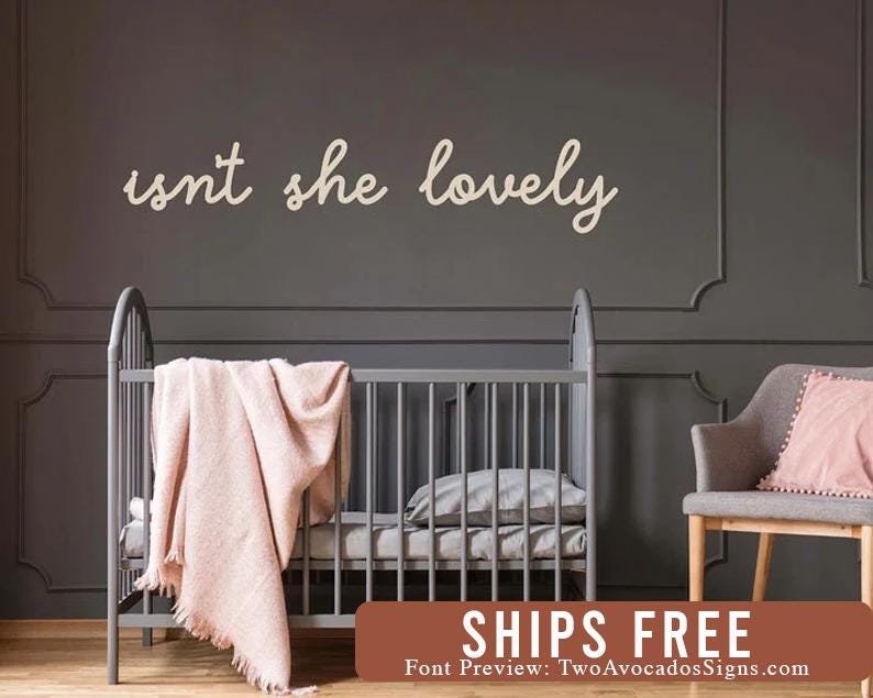 May include: A wall-mounted sign featuring the cursive text "isnt she lovely" in a light cream color. The sign is positioned above a gray crib draped with a soft, light pink textured blanket, set against a dark gray paneled wall.
