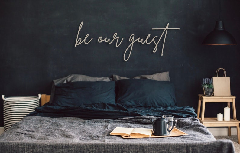 May include: A wall-mounted wooden sign featuring the cursive text be our guest in a light tan finish. The sign is displayed above a bed with dark bedding, a wooden tray holding an open book and a black teapot, a striped laundry basket, and a side table.