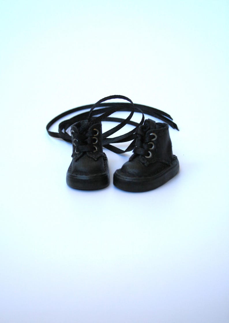 May include: A pair of miniature black leather boots with matching laces. The boots have a classic lace-up design with silver-colored eyelets. The boots are set against a white background.
