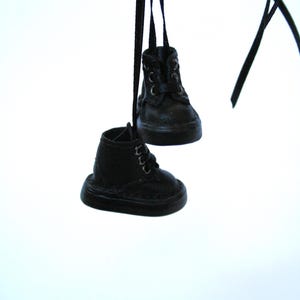 May include: Two miniature black leather boots hanging from black ribbons. The boots have laces and a classic design, with visible stitching details. The background is a plain white.